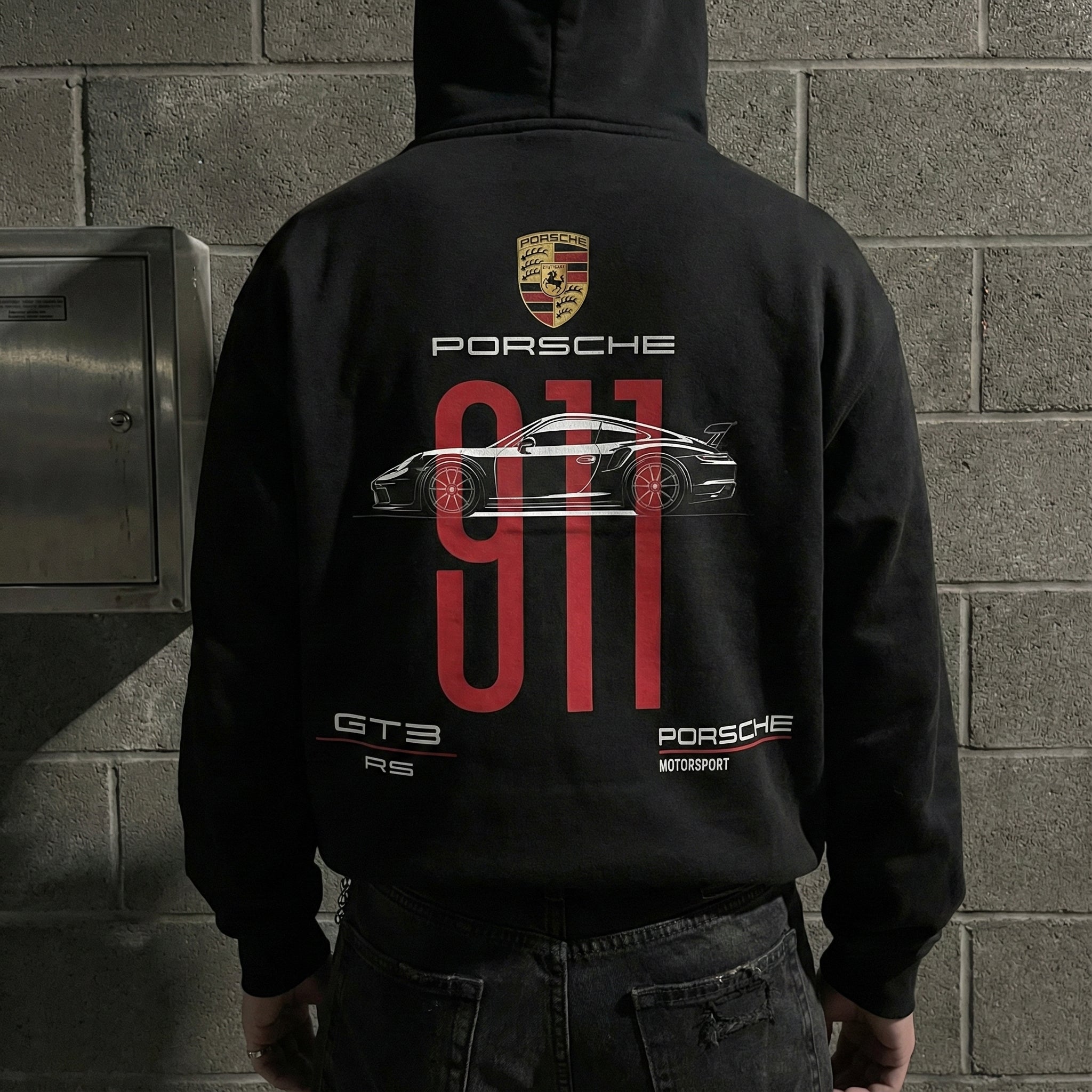Rear view of a person wearing a black hoodie featuring a large graphic design on the back dedicated to the Porsche 911 GT3 RS. The design includes a gold Porsche crest at the top, followed by the text "PORSCHE," and a white side-profile illustration of the car layered over large, vertical red numbers reading "911." The bottom of the design features text reading "GT3 RS" on the left and "PORSCHE MOTORSPORT" on the right. The person is standing in front of a gray cinder block wall.