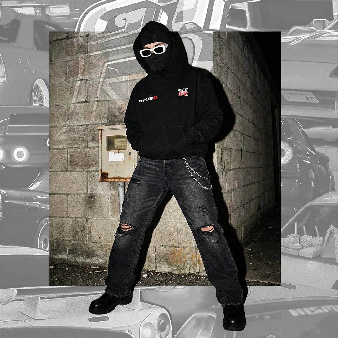 Front view of a model wearing a black hoodie with a face mask and white sunglasses. The hoodie features small, minimalist logos on the chest: "NISMO" on the right and the "GT-R" emblem on the left.
